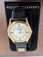 Front view of Caravelle by Bulova wristwatch with gold-tone bezel and black leather strap on black velvet cushion inside box