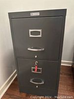 Front view of a gray 2-drawer vertical filing cabinet showing drawer handles, label holders, and lock with key on the lower drawer.