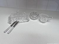 Full set showing the rectangular butter dish with lid, two butter knives with clear handles, cream dish, and sugar dish, all made of clear patterned crystal.
