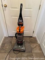 Full front view of upright Hoover WindTunnel Rewind vacuum cleaner showing orange and gray body, black handle, and lower unit on tile floor.