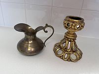 Photo showing both items side by side on a white surface against white tile background, brass pitcher on left, gold Florentine candle holder on right