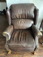 Front view of brown leather recliner chair showing padded cushions, studded armrests, and wooden claw feet on hardwood floor