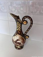 Photo of the front side of the hand painted ceramic pitcher vase showing detailed artwork, curved handle, and gold highlights on a black base.