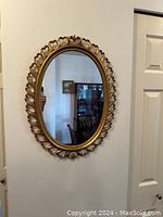 Front-facing photo showing the oval mirror hung on a wall, capturing the detailed gold filigree frame and beaded inner border.