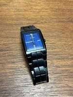 Black Fossil men's wristwatch with blue textured rectangular dial, silver hands, and black metal bracelet laid on wooden surface.