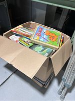 Open cardboard box showing a variety of children's books stacked inside with colorful covers including Bugs and Friends activity book on top.