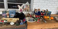 Full overview of the mixed lot items arrayed on two wooden surfaces showing varied collectibles, holiday items, dolls, pet bowls, and candlesticks.