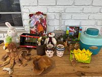 Front view photo showing most items in the lot spread out on a wooden surface with a white brick background, including plush toys, mugs, bowls, and a jug.