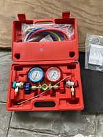 Full view of refrigerant gauge set inside red plastic carrying case, showing blue and red gauges and coiled hoses in top compartment.