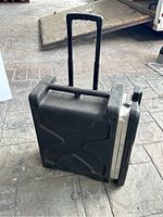 Full view of black plastic hard case with extended telescoping handle and wheels on bottom for transport.
