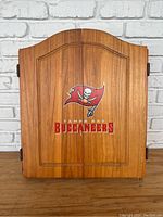 Closed wooden dartboard cabinet with Tampa Bay Buccaneers logo on the front door.