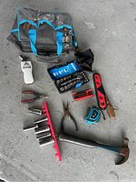 Photo showing tool bag positioned behind various hand tools spread on concrete floor, including hammer, socket set, pliers, tape measure, level, and utility knife.