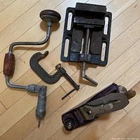 All four vintage hand tools laid out on a wooden floor: a Stanley hand planer, a handle-propelled screwdriver, a metal clamp and a metal table vice.
