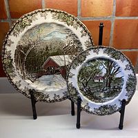 Two Johnson Bros. plates displayed on stands. Larger plate shows a snowy covered bridge scene; smaller plate shows a house and trees scene. Both have decorative green-black borders.