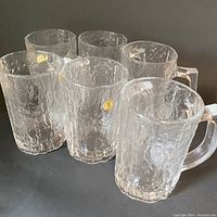 Front and side view of six clear beer mugs with bark-like textured surface and square handles shown arranged together.