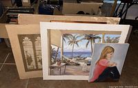 Various vintage and handpainted art pieces, prints, and empty frame arranged leaning against a chair and boxes on tile floor.