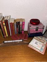 Assorted books, a red heart-shaped digital alarm clock, a decorative pink patterned metal case, pen set in plastic packaging, two red notebooks arranged on a shelf