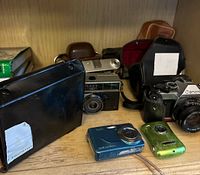 Wide view of the lot showing Canon AE-1 with 50mm lens, vintage film cameras, digital Kodak camera, green metallic pocket camera, and equipment cases.
