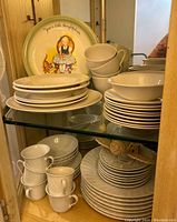 Display of assorted white swirl pattern china plates, bowls, and cups with Hollie Hobby decorative plate standing upright behind.