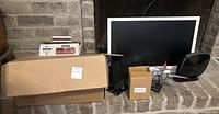 Photo showing a cardboard box with cords and a door chime unit box beside a Vizio TV, Acer speaker, Texaco airplane, and Belkin device all placed in front of a fireplace.