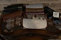 Photo of multiple pairs of metal-framed prescription eyeglasses and several assorted eyeglass cases in various colors and materials arranged on a wooden surface.