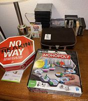 Photo shows a table with a lamp (not included), No Freakin Way card game in a red octagonal box, U-Build Monopoly board game box, stack of black CD cases, cassette tapes, VHS tapes, and a brown fabric vintage cassette carrier case.