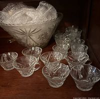 Photo of large clear etched glass punch bowl with matching etched cups and additional cups with different design in a wooden cabinet shelf.