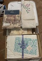 Photo of assorted linens including boxed lace tablecloth, embroidered linen cloth, and packaged floral fabric