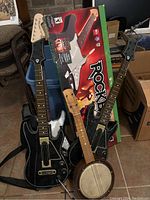Three musical instruments grouped together on floor — two Rock Band guitar controllers and one wooden banjo.