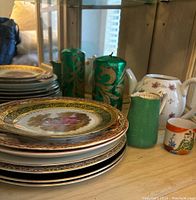 Stack of ornate decorative plates with gold trim and detailed painted scenes, with green cylindrical candles and porcelain mini cup and teapot in background