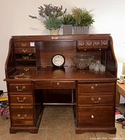 Full front view of the wood roll top desk showing the roll top open, multiple small storage cubbies, and drawers below with brass hardware. The desk is dark wood with signs of wear and peeling on the top surface. Decorative items are placed on top.