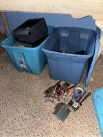 Two blue plastic totes under a wooden table with assorted tools and a black paint sprayer in one tote. Various paint brushes, putty knives, and a roll of blue painter's tape placed on carpeted floor in front of totes.