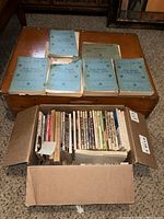 Box containing various vintage books and magazines in worn condition on a wooden chest.