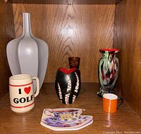 Wide view of vintage collectibles including frosted bottleneck vase, handpainted Sarn vase, swirled glass vase, dark glass candleholder, golf mug, mini orange tea cup and tea notepad