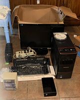 Opened cardboard box containing Lenovo desktop tower, flat-panel monitor, keyboards, mouse, power strip, external hard drive, data tapes, and cables arranged on the floor.