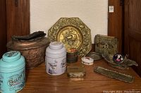 Photo showing various tins including English Tea tins, rustic copper planter, and brass wall plaque with animal motif, arranged on wooden shelf.