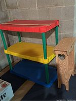 Three-tier children's plastic shelf with red, yellow, and blue tiers and green supports next to a wooden shelf with heart cut-out sides.
