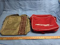 Photo showing side-by-side front view of two TWA vintage travel bags on a blue surface with a ruler in front for scale.