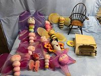 Wide shot showing multiple colorful Flutter Friends plush decorations including caterpillars, bees, and a butterfly, along with a miniature wooden chair and a small piano with box on a light blue backdrop.