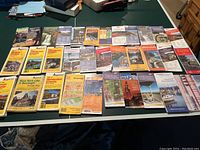 Top view of approximately 30 road maps of US states, Canada, and international locations arranged in rows on a table. Booklet and fold-out style maps with colorful covers.