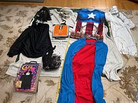 Photo showing a variety of Halloween costumes laid out on a blanket, including superhero themed costume shirt, multiple capes, and masks such as Darth Vader and skull faces