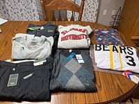 Overview of men's clothing lot with seven items including slacks, tees, sweaters, jerseys, and hoodies shown spread on a table with wooden chair background.