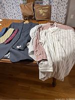 Photo of men's navy suit jacket alongside red plaid and striped dress shirts with three ties and garment bag visible in background.