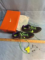 Pair of black Nike track shoes with neon green laces and purple interior, placed next to an orange Nike shoe box and measuring stick, photographed to show overall condition and included spikes.