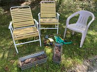 Three outdoor chairs: two white metal fold-up chairs with yellow slats and one beige resin chair. Two rectangular metal deck railing potted planters and assorted outdoor watering supplies including hose and nozzles are positioned on grass outdoors.