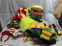 Assorted colorful party hats including a large green furry hat, red and white striped hat, foam beer mug hat, and jester hats alongside Mardi Gras bead necklaces and various gift bags.