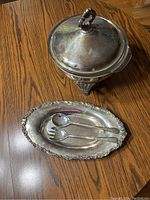 Silver plated chafing dish with lid, silver plated serving dish, and two serving utensils on wooden table.