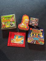 Top-down photo of various nostalgic game and book items including Nickelodeon Scene It DVD game, Simpsons CD, Homer shaped toy, puzzle box, and Marvel Family Feud.