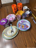 Photo showing the full assortment of toddler dishware items including divided plates, bowls, cups, mugs, tumblers, and utensils arranged on wood table.