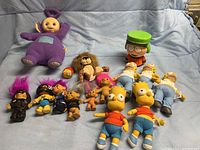 Shows the full lot of collectible plush toys and figurines including Teletubbies, Simpsons, Trolls, South Park, Madagascar, and Curious George characters on a blue blanket.